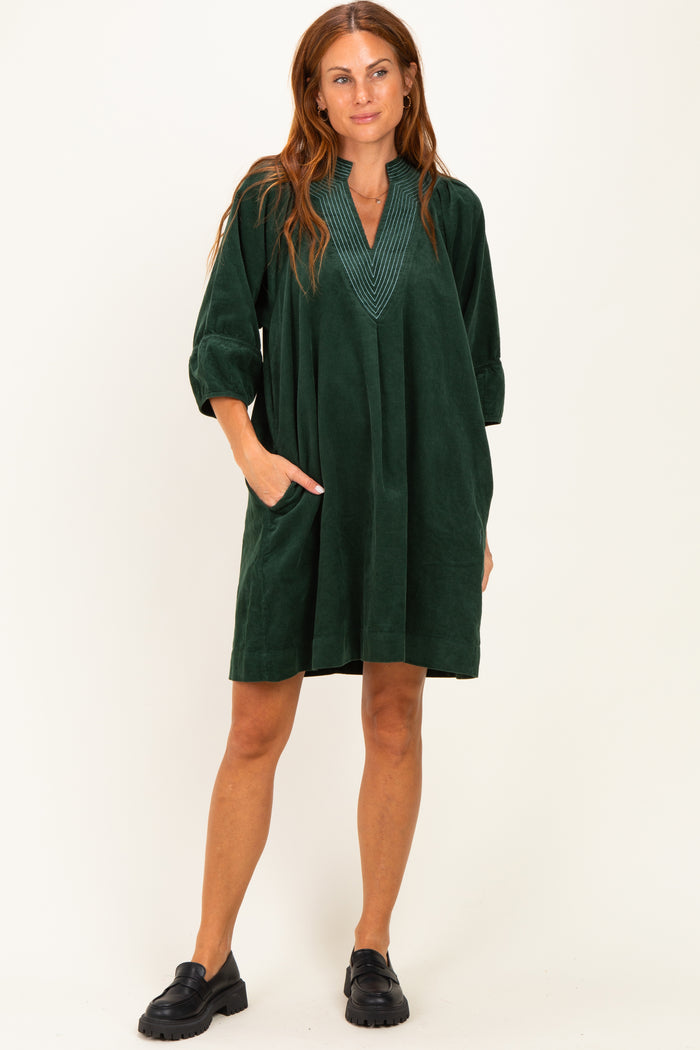 woman wearing a dark green corduroy tunic dress with a v-neck and pockets, styled with black slip-on shoes