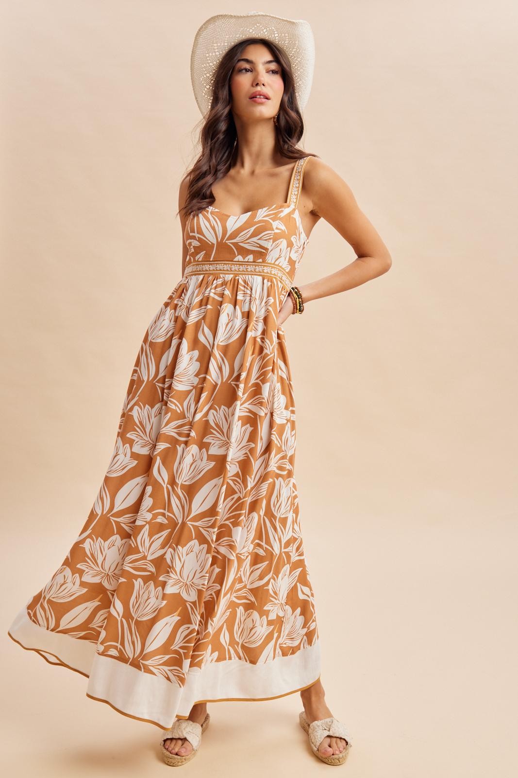 woman wearing an orange floral maxi dress with a square neckline and straps, posing stylishly with a wide-brimmed hat
