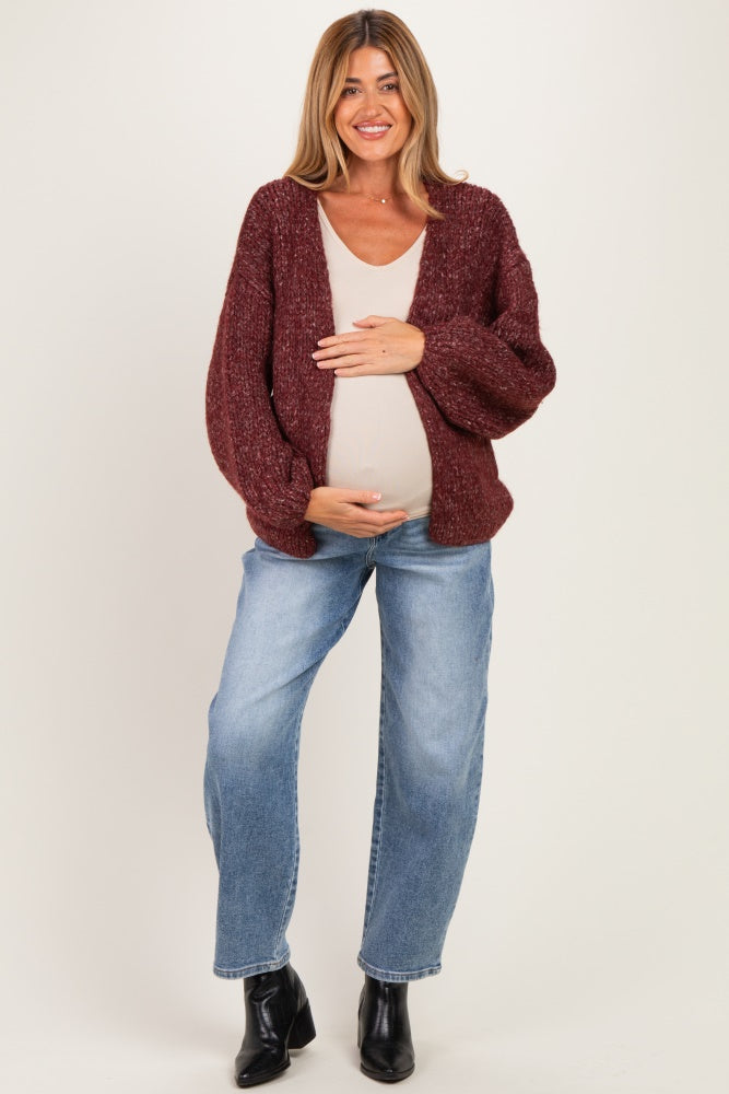 woman wearing a burgundy chunky knit maternity cardigan and light tank top, cradling her baby bump while standing in jeans