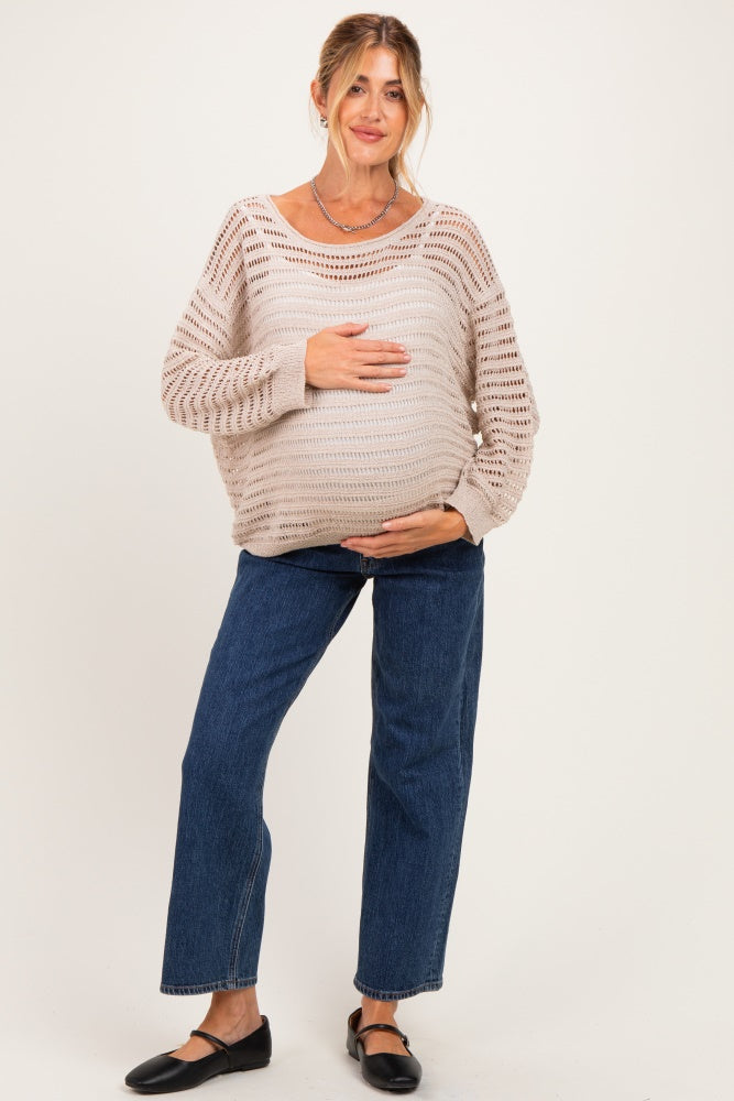 woman wearing a beige open-knit maternity sweater and blue straight-leg jeans, holding her baby bump