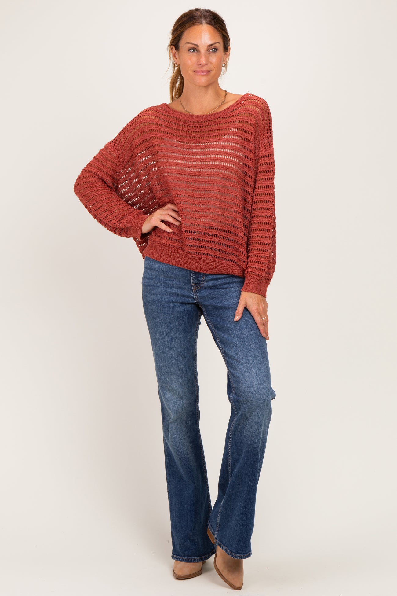 woman wearing a rust-colored open knit sweater and flared jeans, posing with one hand on her hip