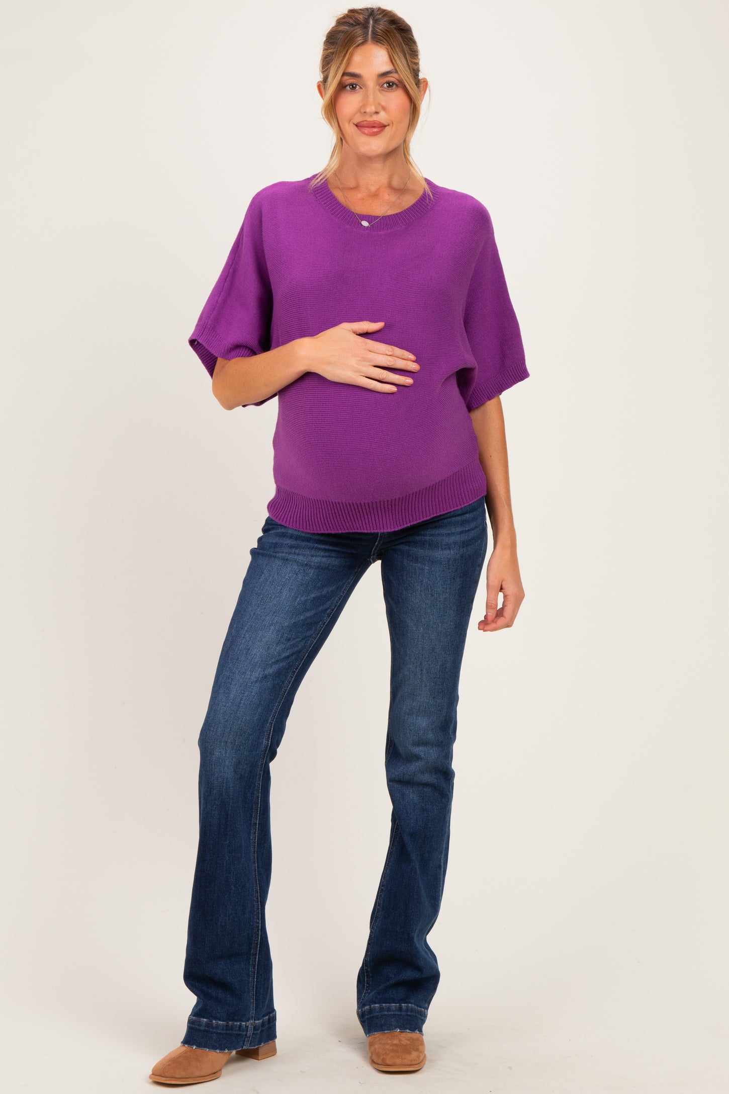 woman wearing a purple maternity sweater and blue flared jeans, gently cradling her baby bump