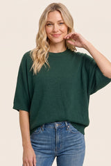 woman wearing a dark green oversized sweater and light blue jeans, smiling and posing with one hand on her cheek