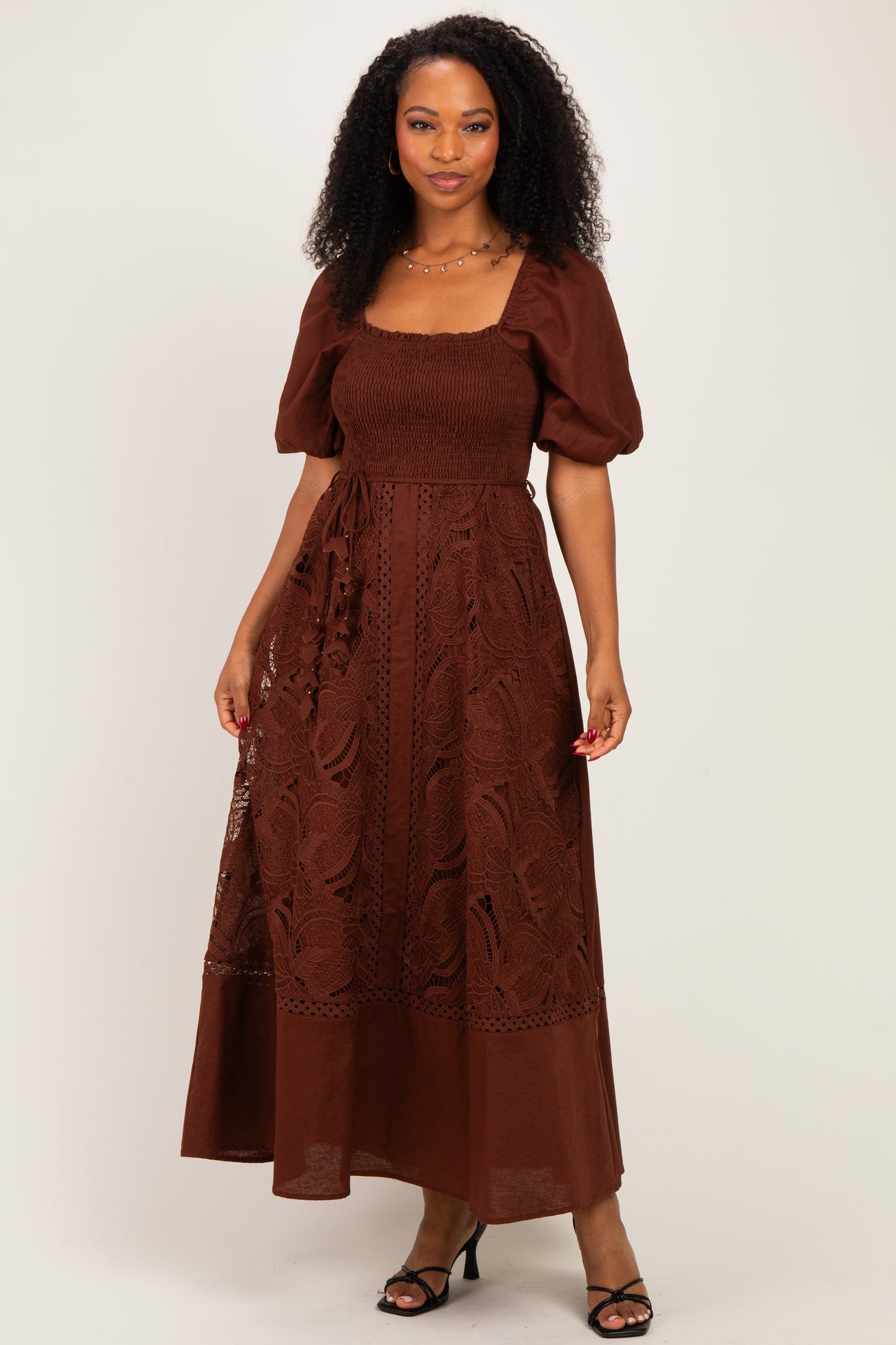 woman wearing a brown maxi dress with puff sleeves and lace detailing, standing with one hand on her hip