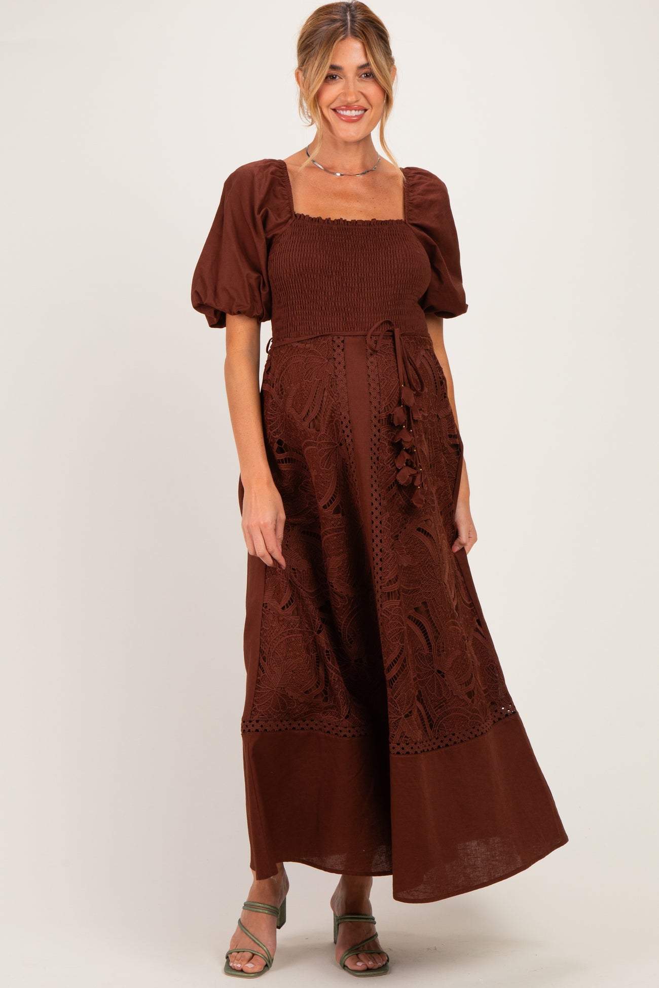 woman wearing a brown smocked maternity maxi dress with puff sleeves and lace detailing, showcasing her baby bump