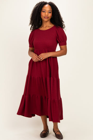 woman wearing a burgundy tiered maxi dress with puff sleeves and a textured fabric, standing against a plain background