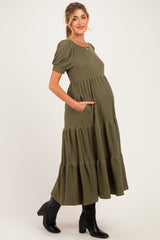 woman wearing a dark olive green maternity tiered maxi dress with short puff sleeves and pockets, showing her baby bump