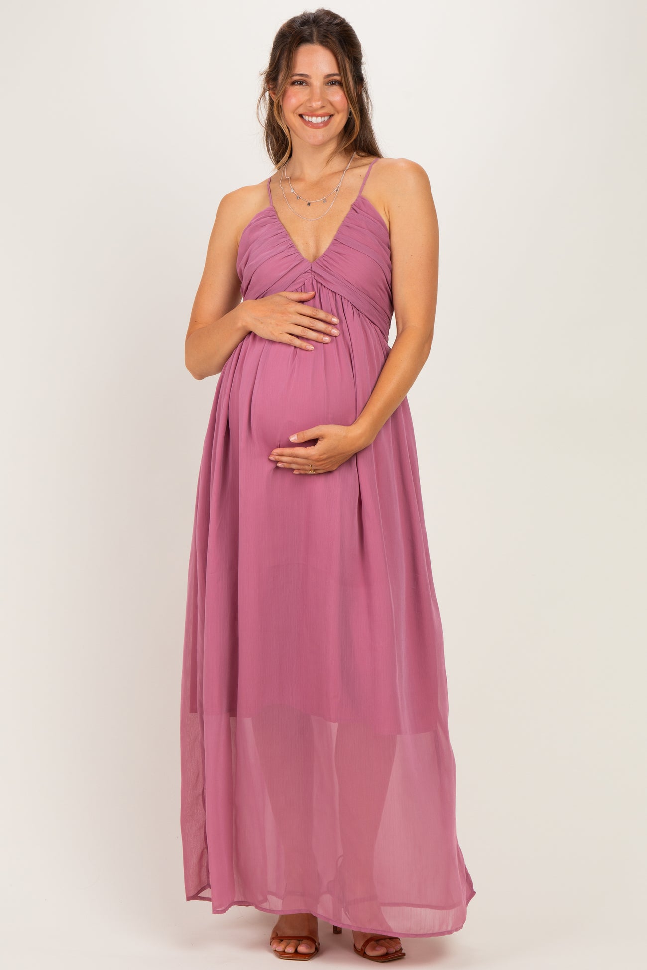 woman wearing a mauve maternity maxi dress with a ruched bodice and sheer overlay, holding her baby bump