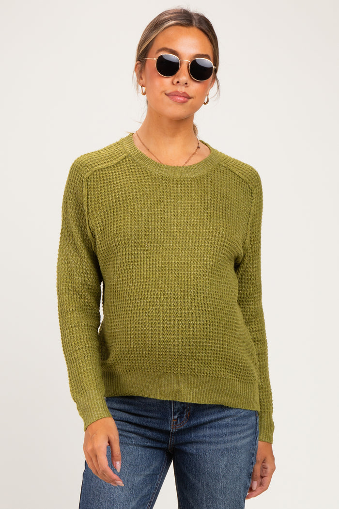 woman wearing a green knit maternity sweater and blue jeans, with a baby bump visible and sunglasses on