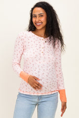 woman wearing a light pink floral maternity top with button details and orange cuffs, holding her baby bump
