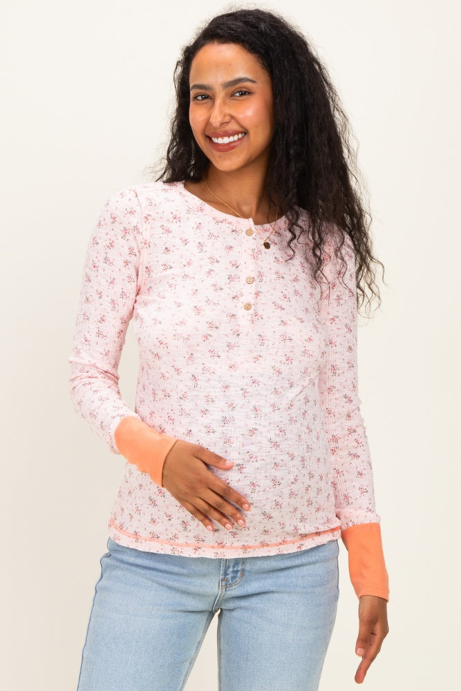 woman wearing a light pink floral maternity top with button details and orange cuffs, holding her baby bump