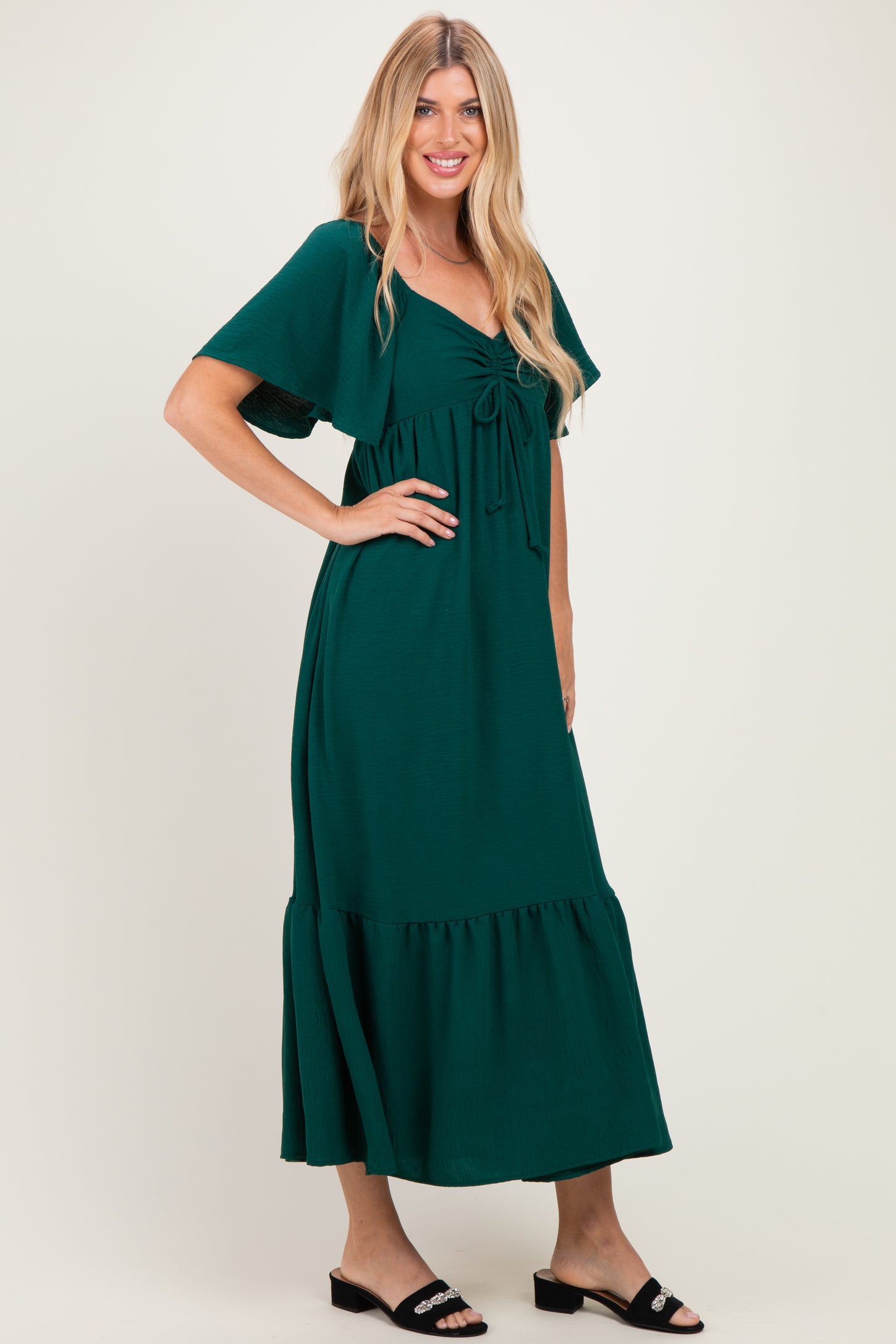 woman wearing a dark green maxi dress with a tie front and flutter sleeves, standing against a light background