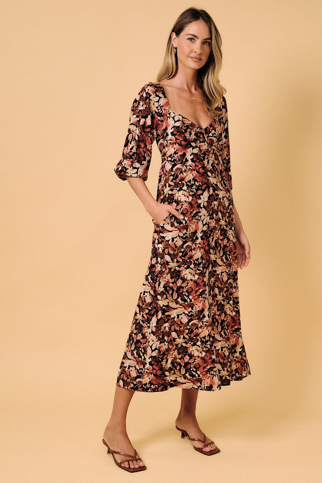 woman wearing a floral midi dress with a scooped neckline and three-quarter sleeves, standing against a beige background