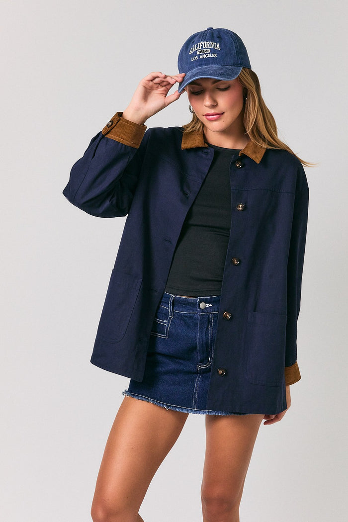 woman wearing a navy blue oversized jacket with a brown collar and a black top, paired with denim shorts and a blue cap
