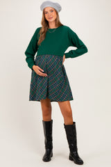 woman wearing a green long sleeve maternity dress with a plaid skirt and holding her baby bump