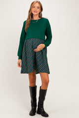 woman wearing a green maternity dress with a plaid skirt and long sleeves, holding her baby bump