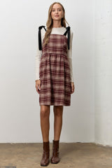 woman wearing a burgundy plaid dress with black tie straps and a cream long sleeve top, standing on a concrete floor
