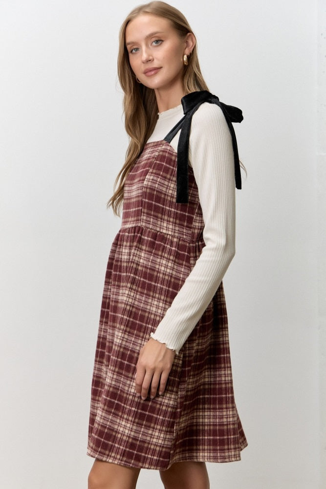 woman wearing a maroon plaid pinafore dress over a white long sleeve top with black ribbon details