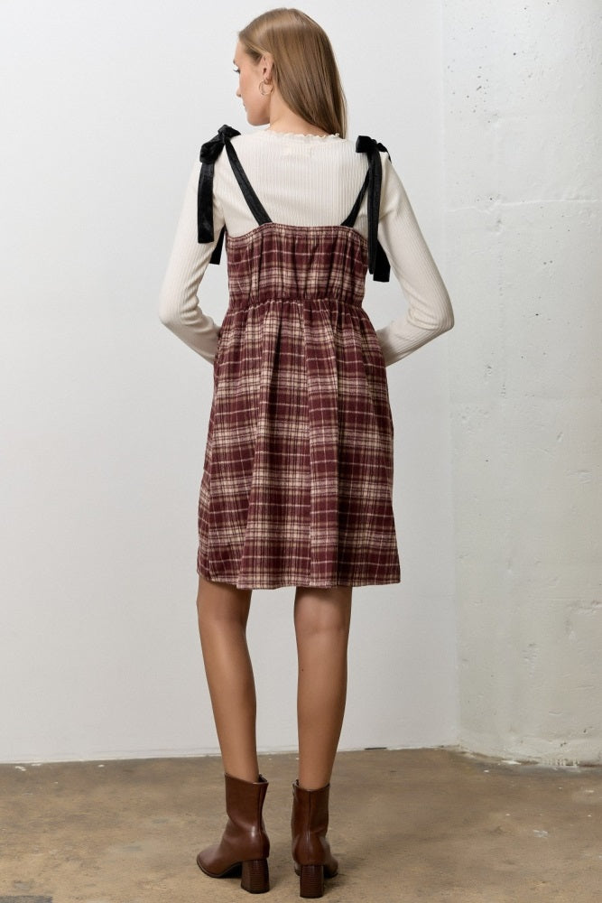 woman wearing a burgundy plaid pinafore dress with black tie straps over a white long sleeve top and brown ankle boots