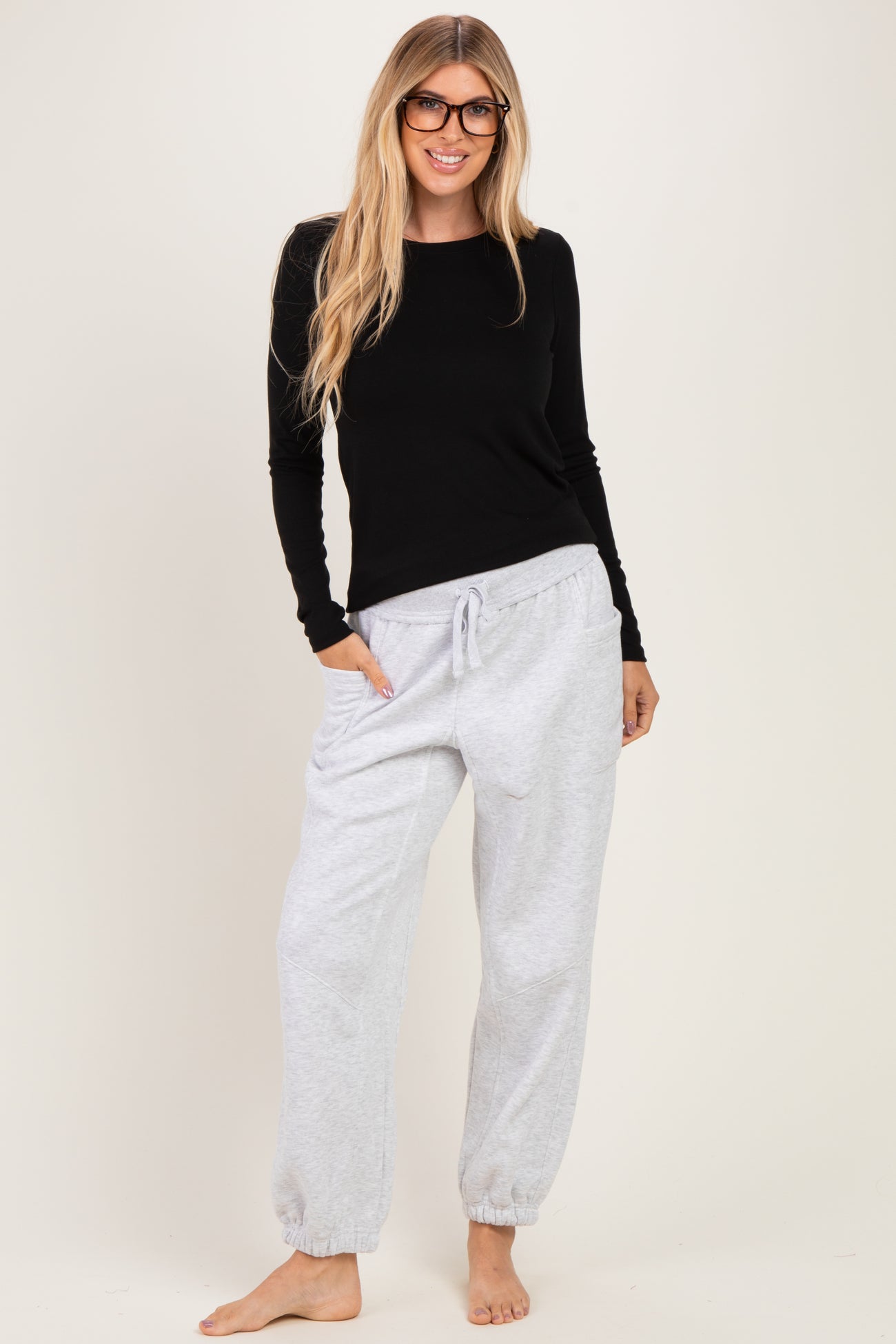 woman wearing a black long sleeve top and light grey drawstring jogger pants, smiling and standing barefoot