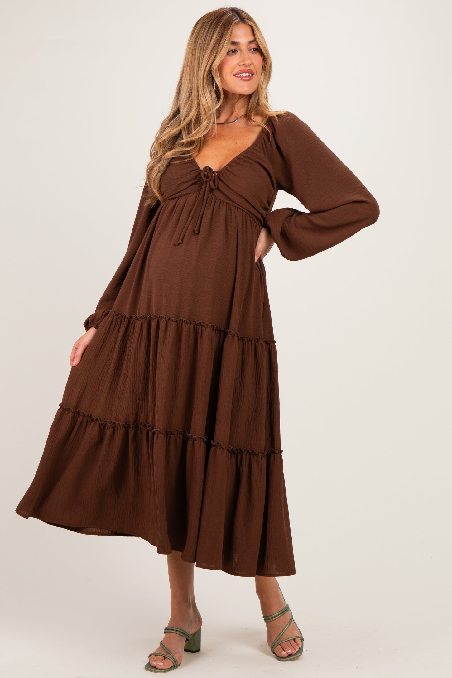 woman wearing a brown maternity maxi dress with a tie front and tiered ruffle details, holding her baby bump