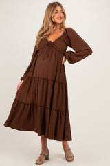 woman wearing a brown maternity maxi dress with a tie front and tiered ruffle details, holding her baby bump