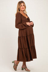woman wearing a brown maternity maxi dress with long sleeves and tiered ruffles, holding her baby bump