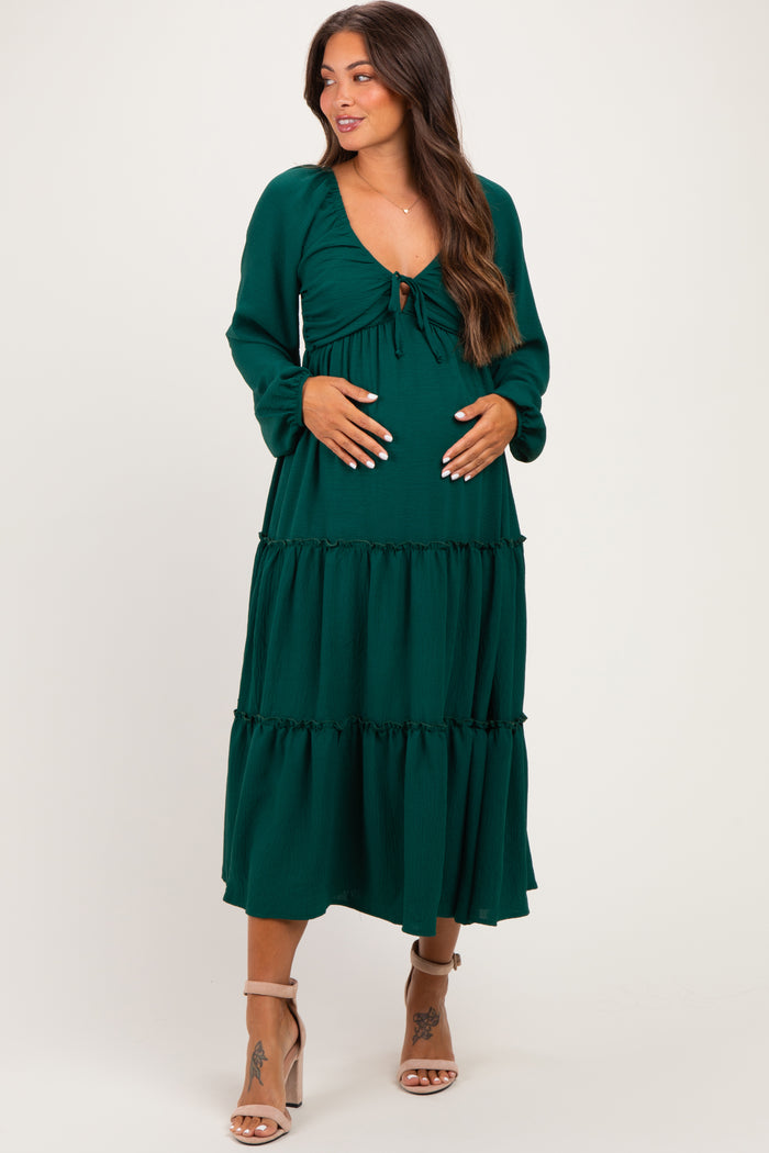 woman wearing a dark green maternity dress with a tie front and tiered skirt, holding her baby bump