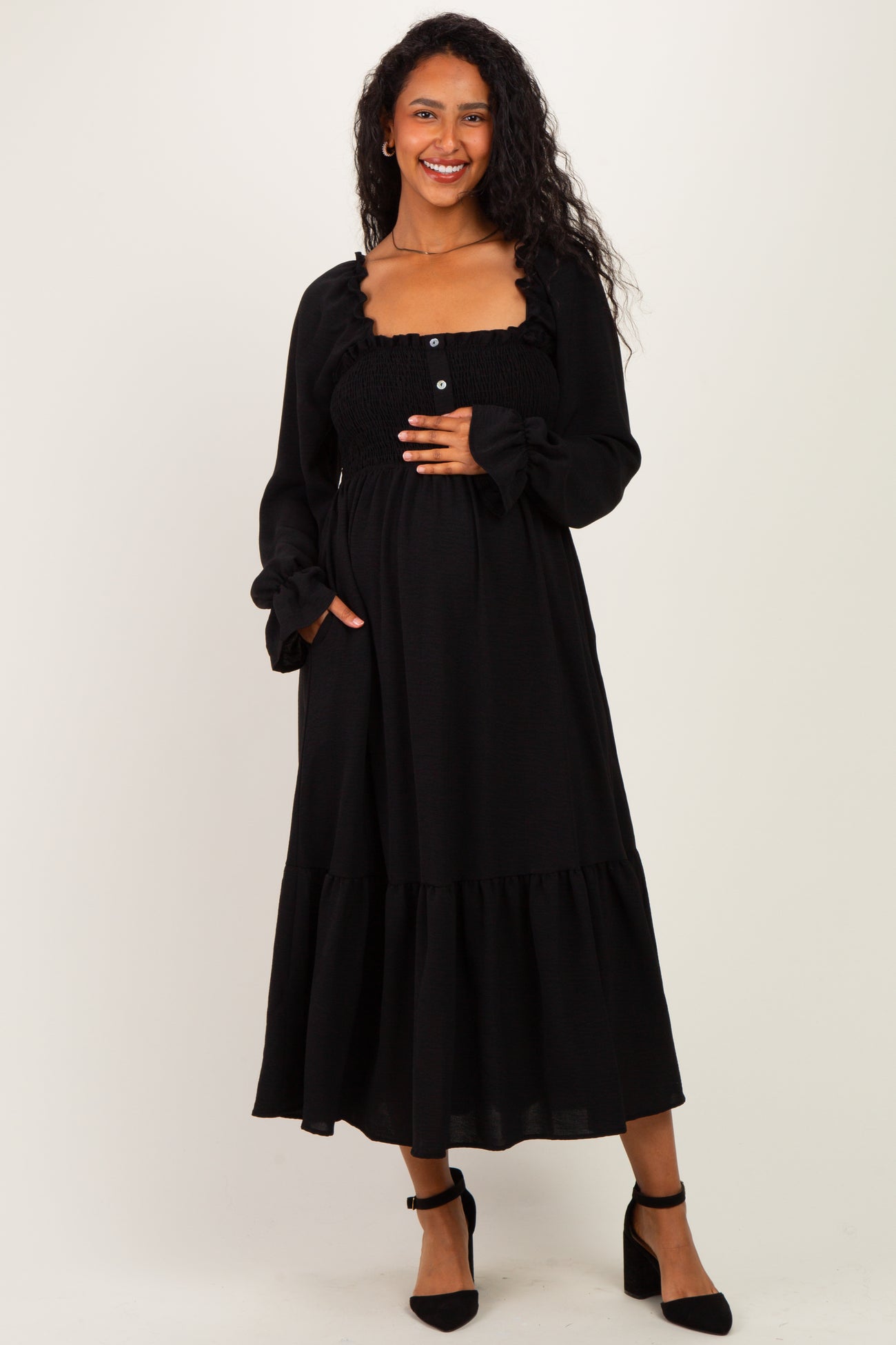 woman wearing a black maternity maxi dress with smocked bodice and ruffled sleeves, smiling and displaying her baby bump