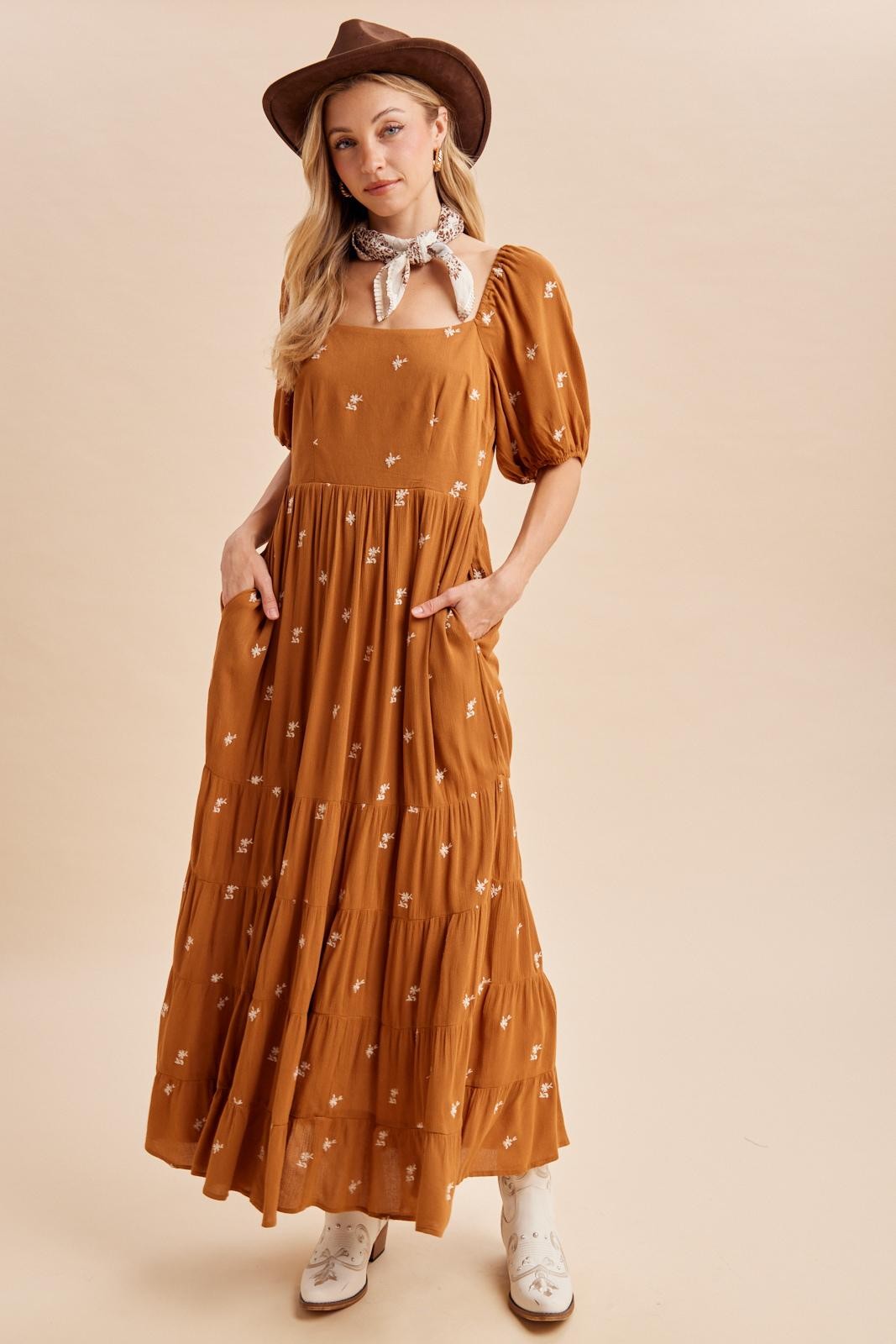 woman wearing a rust colored maxi dress with floral embroidery, puff sleeves, and pockets, styled with a brown hat and scarf