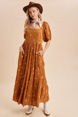 woman wearing a rust colored maxi dress with floral embroidery, puff sleeves, and pockets, styled with a brown hat and scarf