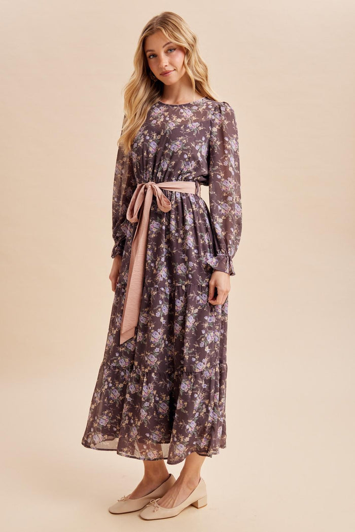 woman wearing a long floral maxi dress with long sleeves and a belt, standing against a neutral background