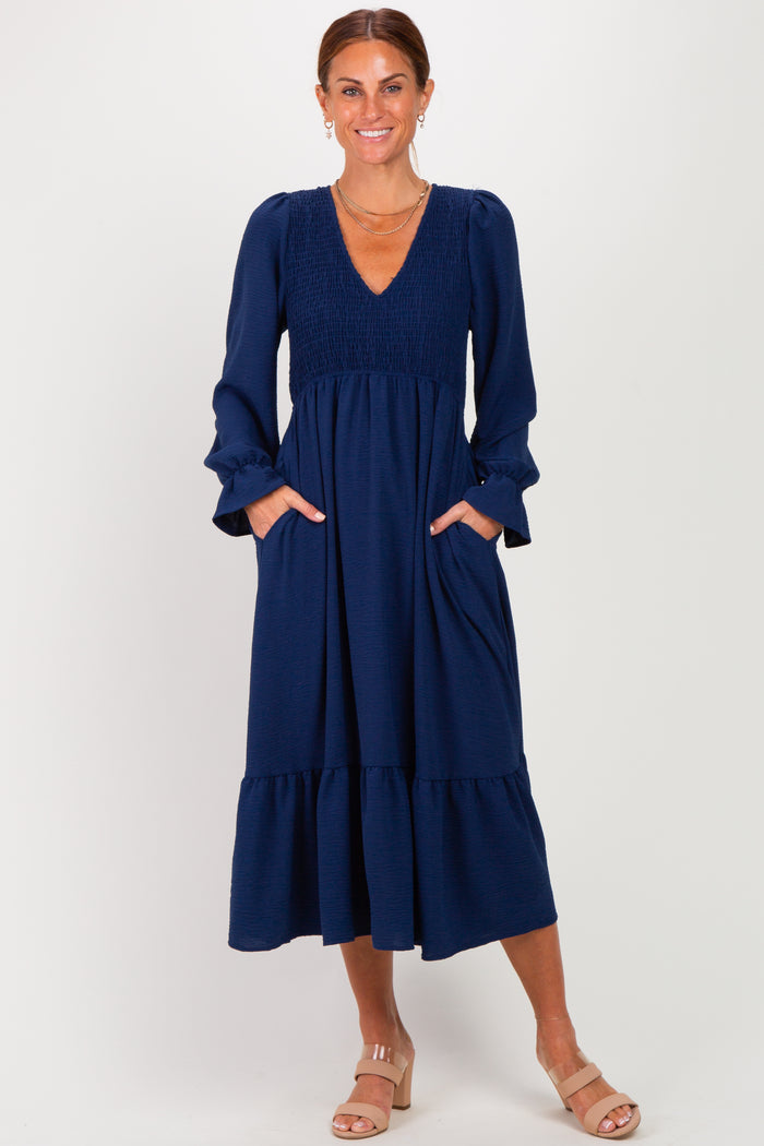 woman wearing a navy blue long sleeve maxi dress with smocked bodice and pockets, standing with hands in pockets