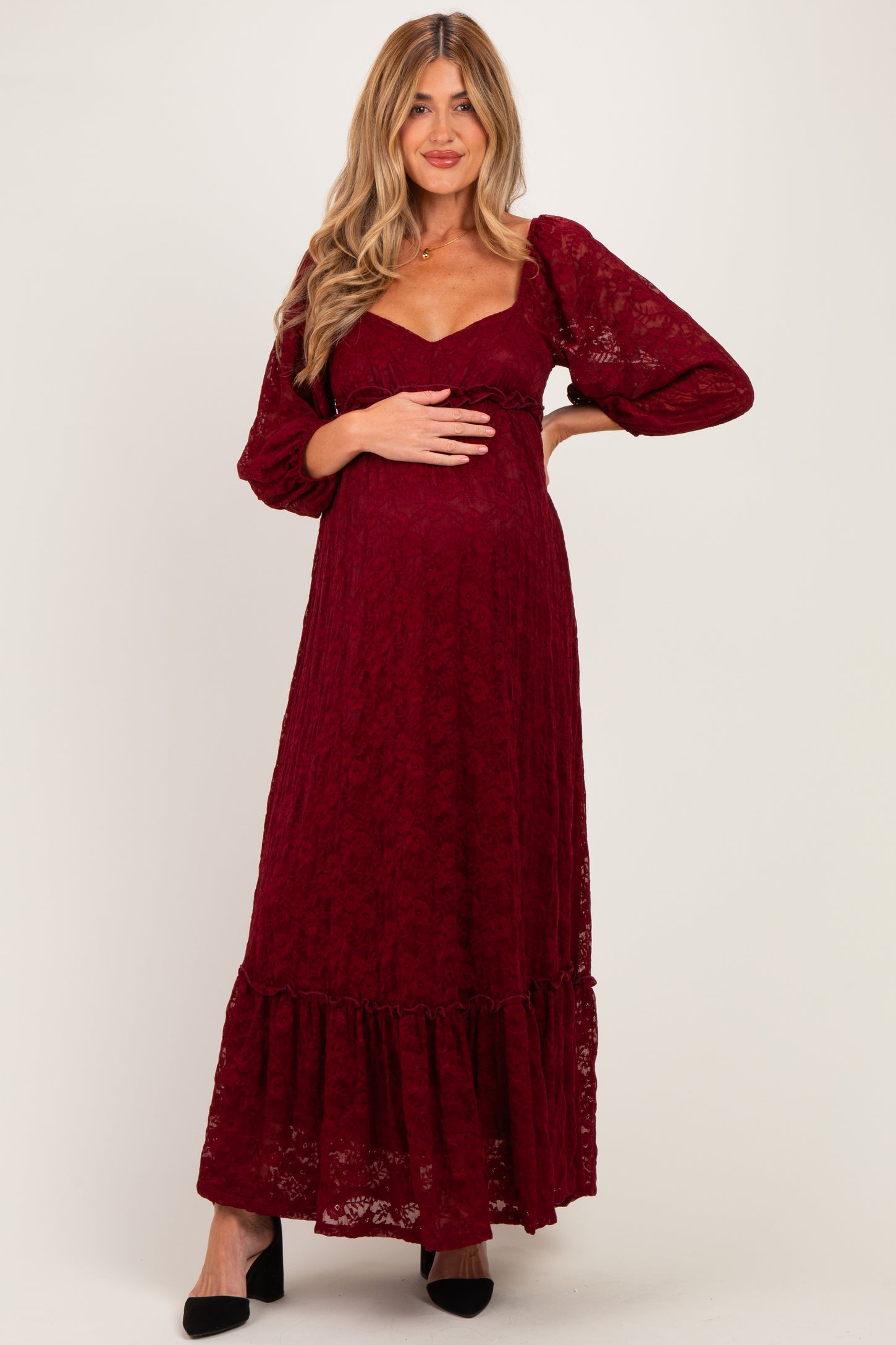 woman wearing a burgundy lace maternity maxi dress with puff sleeves and a ruffled hem, holding her baby bump