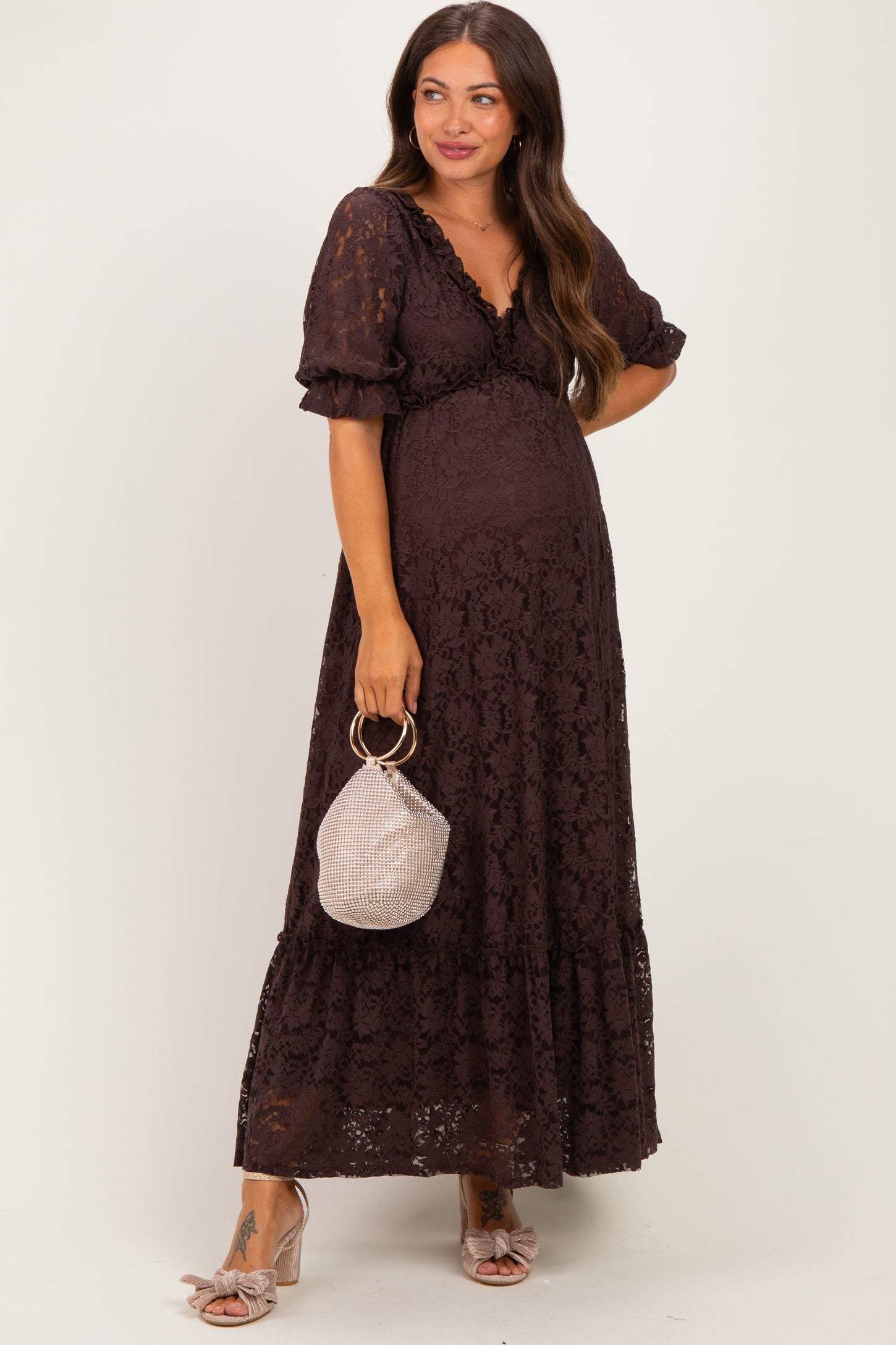 woman wearing a brown lace maternity dress with puff sleeves and a flowy skirt, holding a small handbag and showing her baby bump