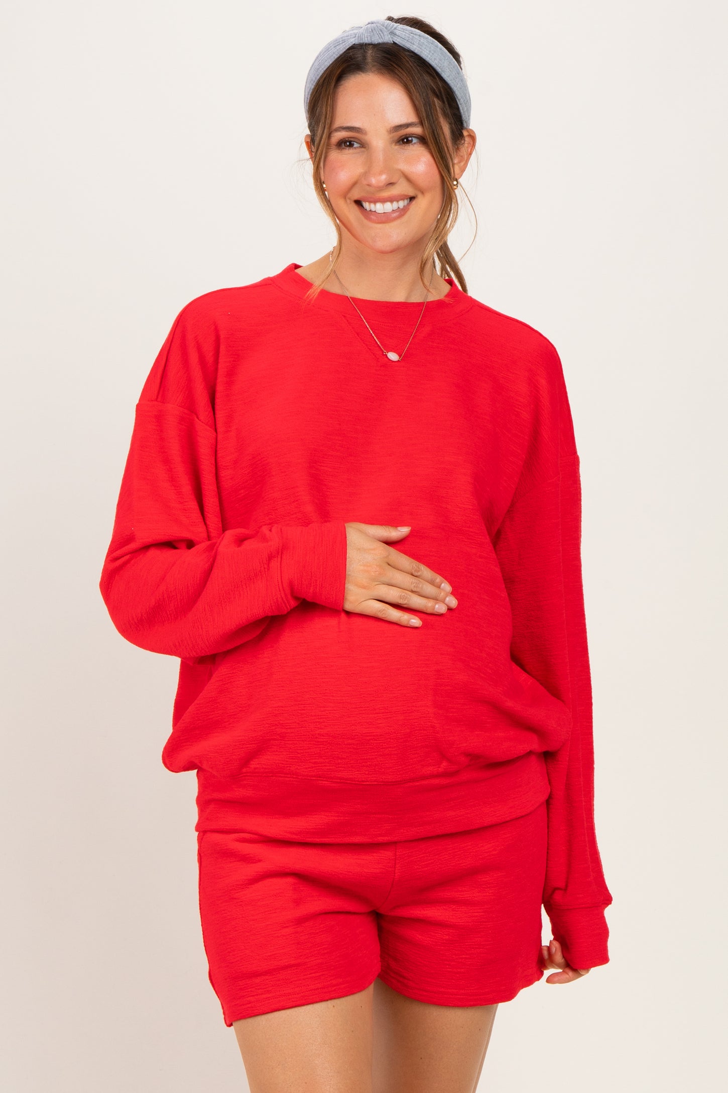 woman wearing a red oversized maternity sweatshirt and matching shorts, smiling and holding her baby bump