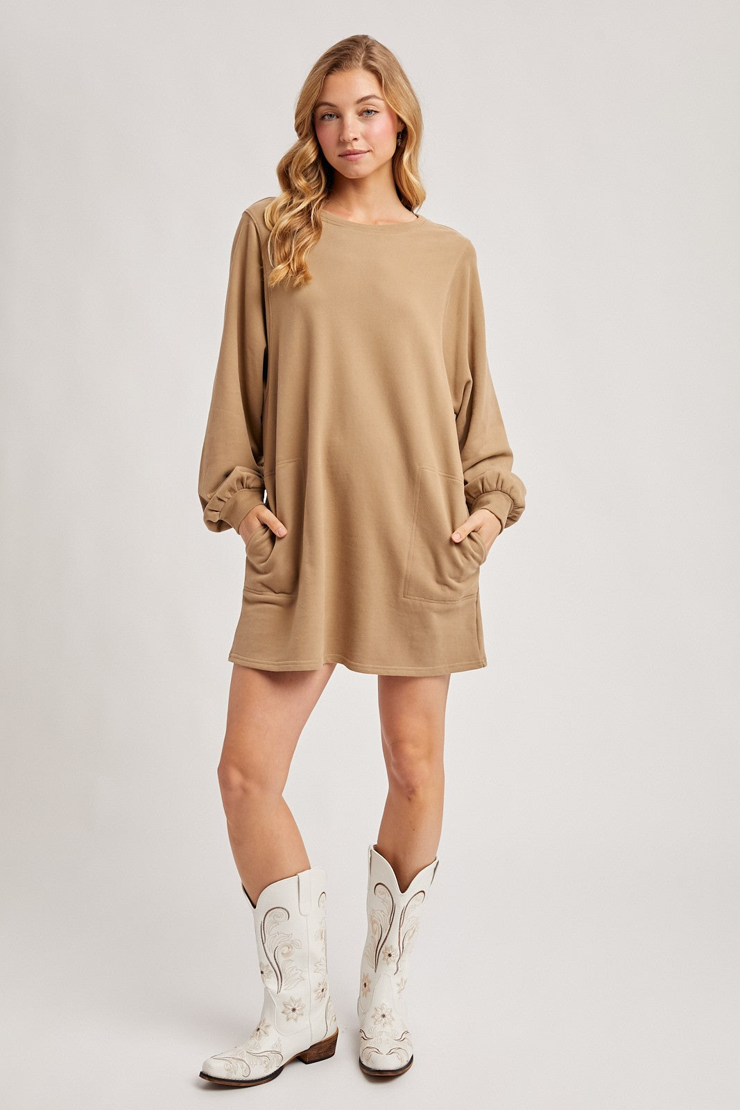 woman wearing a tan long sleeve oversized dress with pockets and white cowboy boots