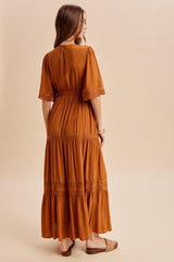 woman wearing a rust-colored maxi dress with lace details and a tiered hem, viewed from the back