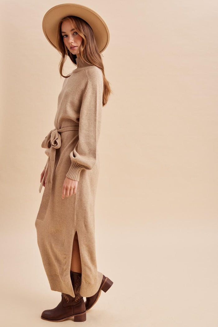 woman wearing a beige long sleeve sweater dress with side slits and a tied waist, paired with a wide-brimmed hat and ankle boots