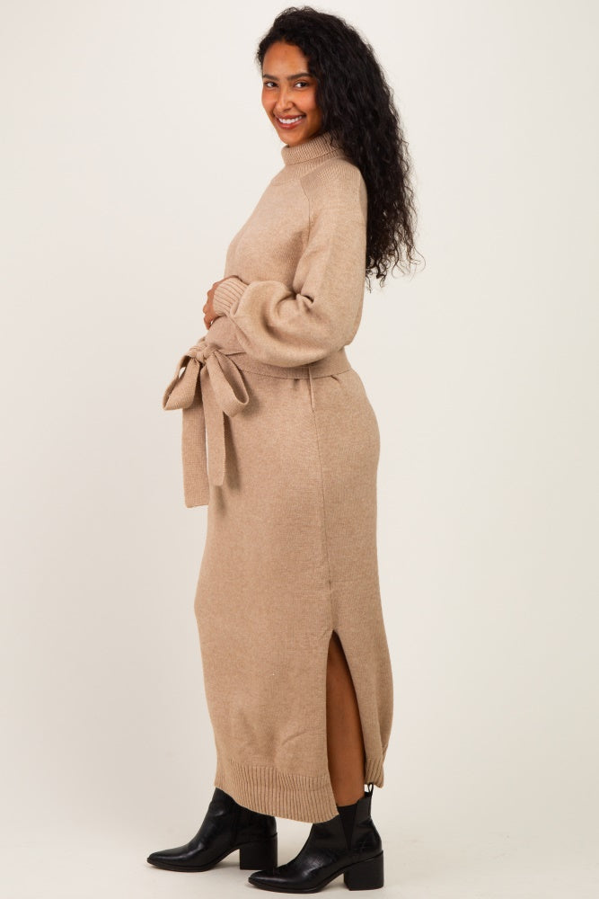 woman wearing a beige maternity sweater dress with a tie waist and side slit, holding her baby bump