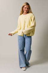 woman wearing a yellow fluffy cardigan over a white top and blue wide leg jeans, smiling while posing against a neutral background