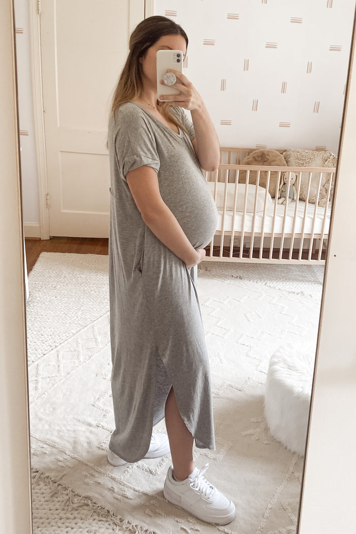 pregnant woman in grey maxi dress taking mirror selfie in a nursery with a wooden crib and white rug