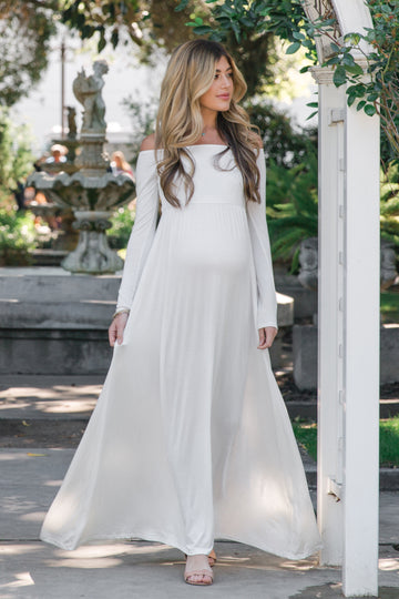 woman wearing a white off-shoulder maternity gown and holding her baby bump in a bedroom setting
