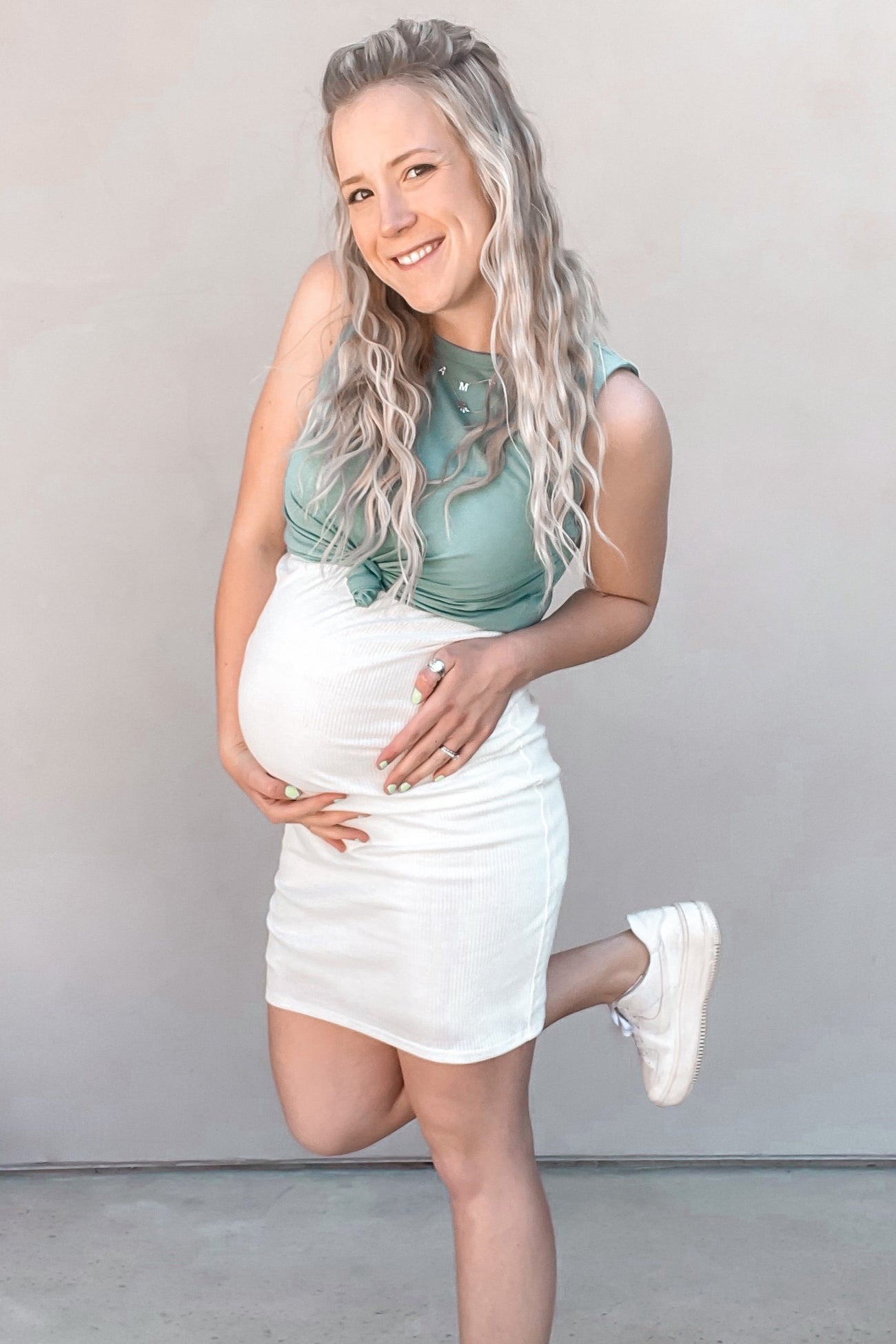 woman wearing a light green maternity tank top and white maternity mini skirt, cradling her baby bump