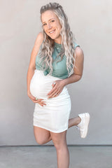 woman wearing a light green maternity tank top and white maternity mini skirt, cradling her baby bump