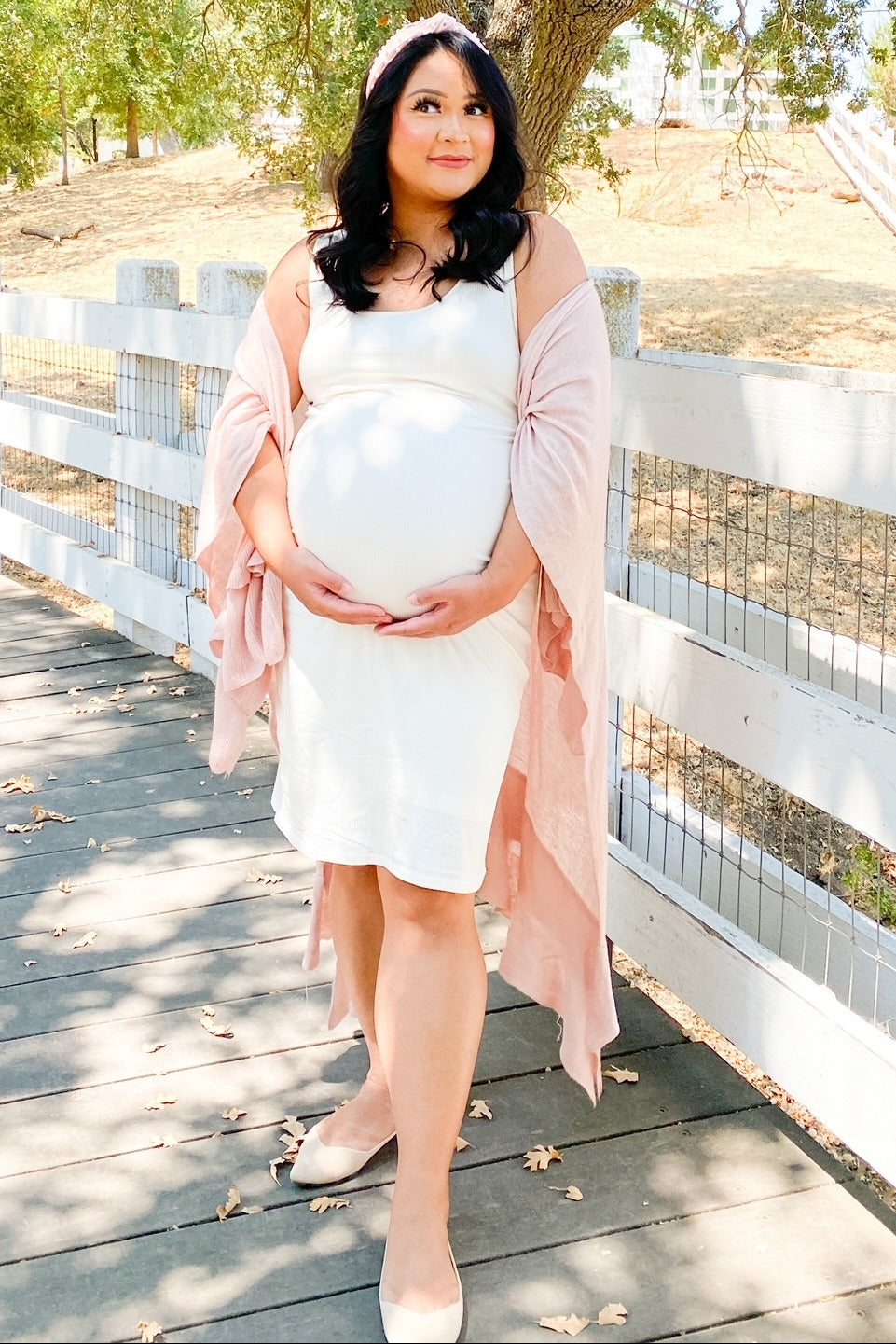 woman wearing a white maternity dress and a light pink shawl, cradling her baby bump outdoors