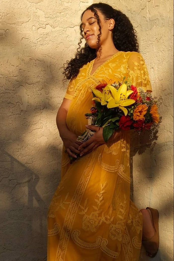 woman wearing a yellow embroidered maternity dress and holding a bouquet of flowers, showcasing her baby bump