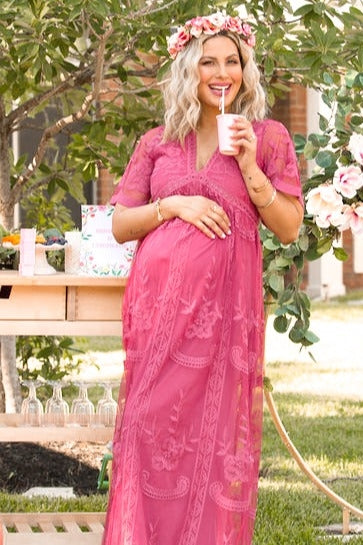 woman wearing a pink embroidered maternity maxi dress, holding a drink and showing her baby bump with a floral crown on her head