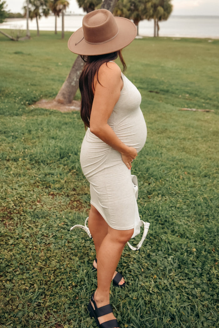 woman wearing a light gray form-fitting maternity dress, showing her baby bump, accessorized with a wide-brimmed hat