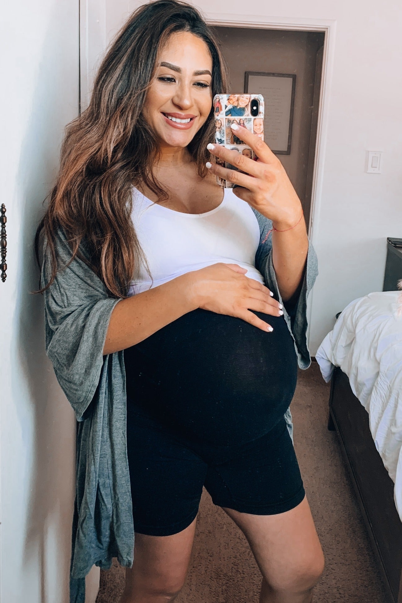 woman wearing a white tank top and black shorts, holding her baby bump, with a gray cardigan draped over her shoulders